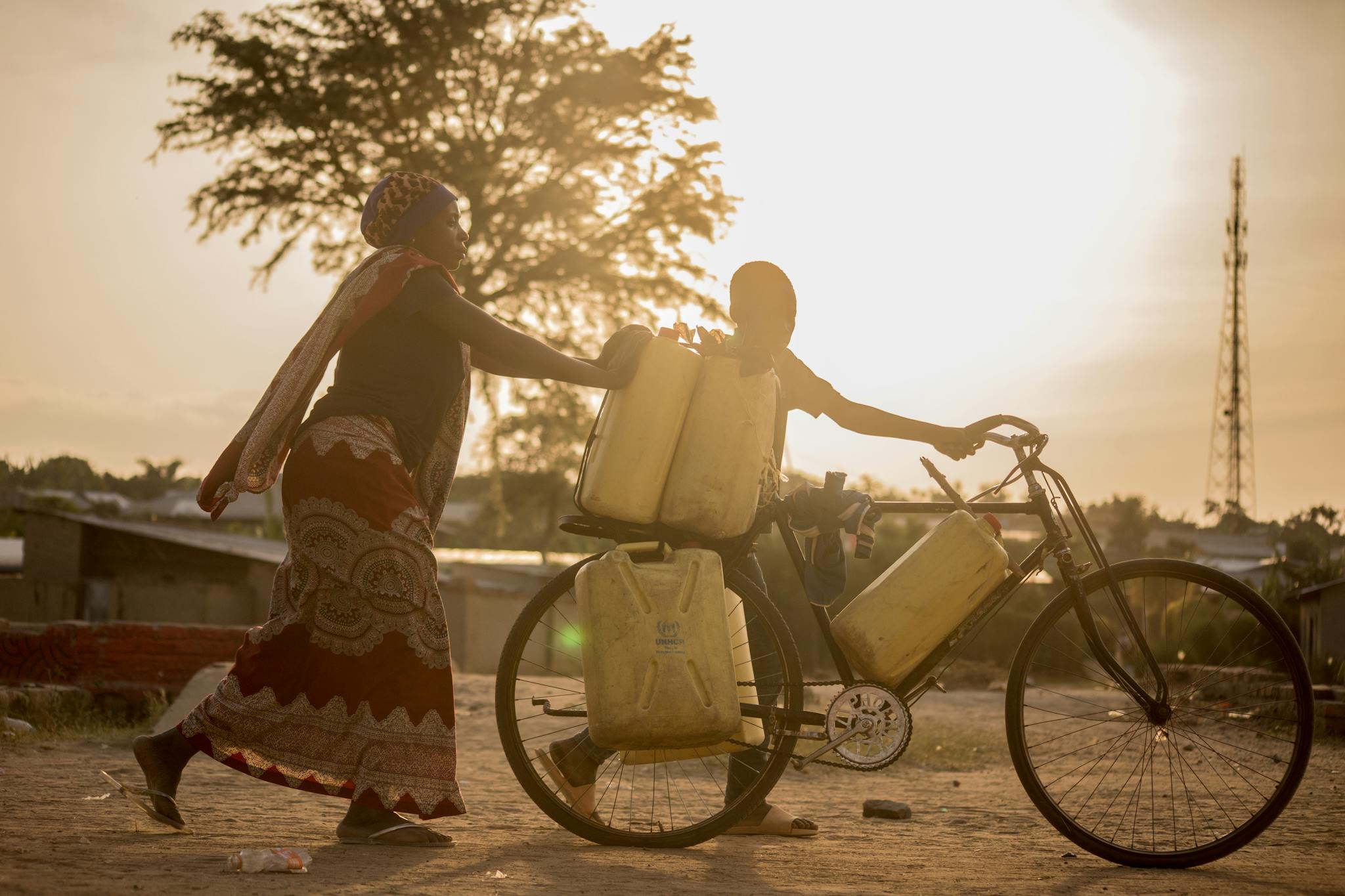 A mother and son move water containers on a bicycle during sunset, Africa.