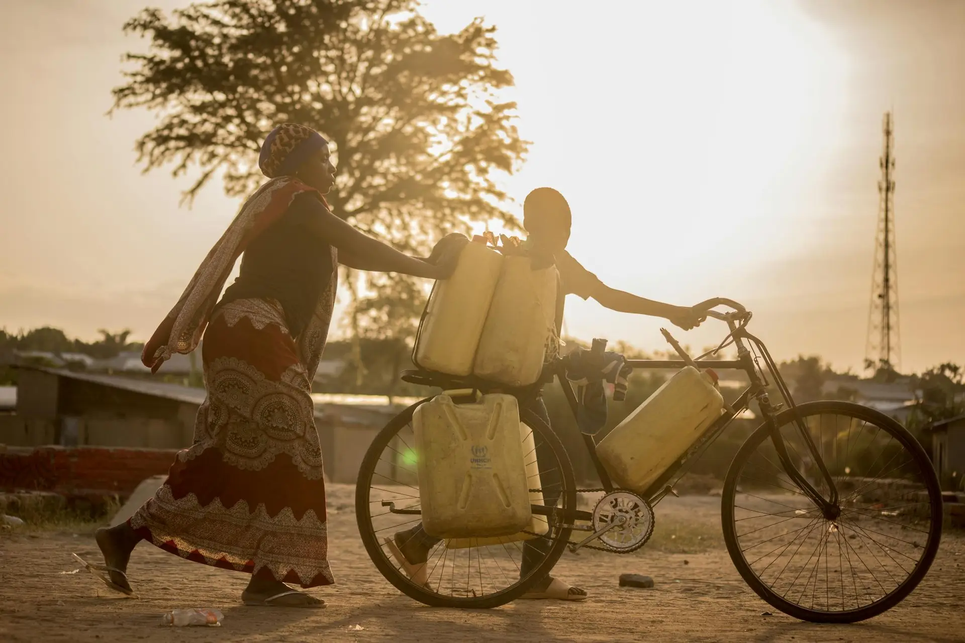 A mother and son move water containers on a bicycle during sunset, Africa.